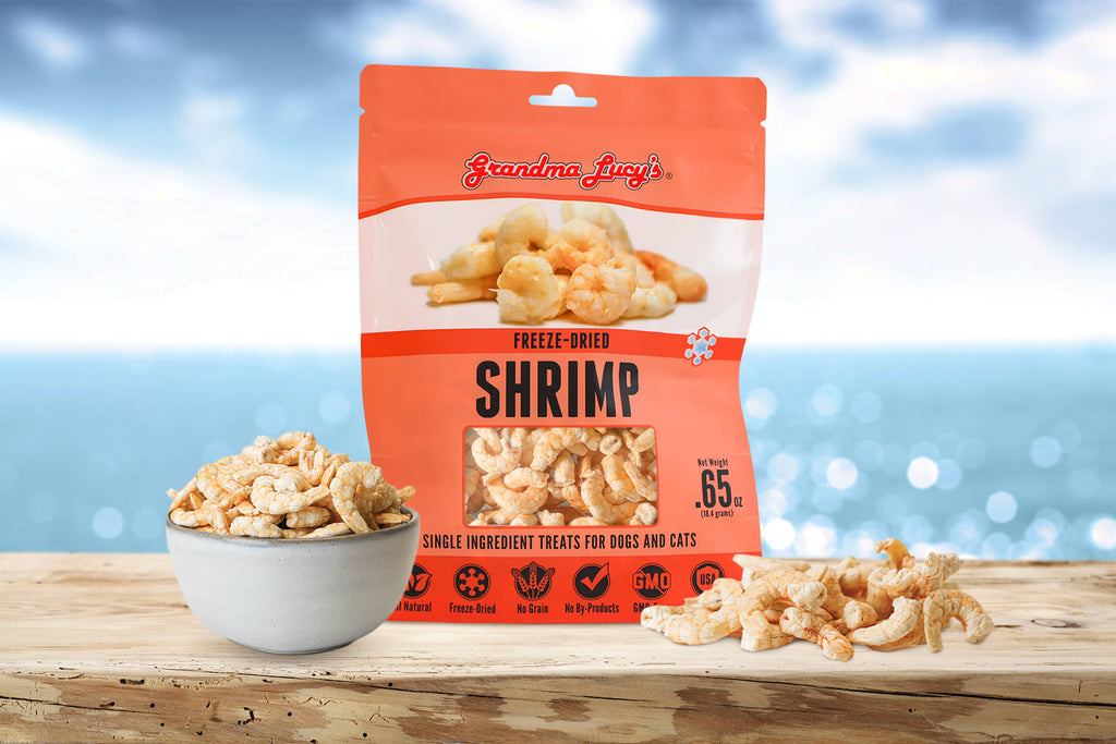 Grandma Lucy's shrimp cat and dog singles treat bag next to a bowl of treats, displayed on a wooden surface with a calming ocean background. 